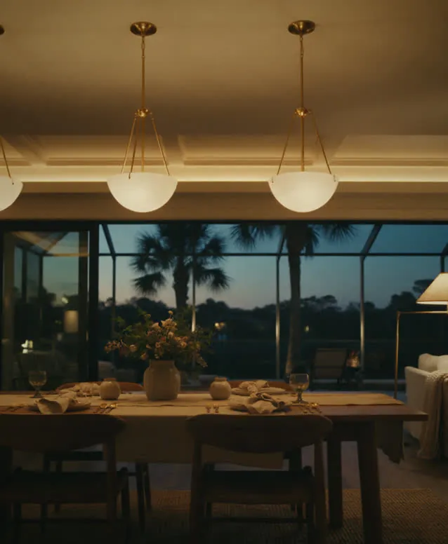 Dining room with warm-dimmed pendants and cove light, lanai and palms beyond the glass